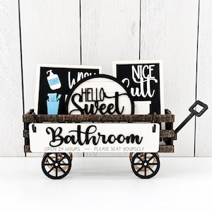 May include: A miniature dark brown wooden wagon featuring a white sign that reads Bathroom OPEN 24 HOURS — PLEASE SEAT YOURSELF. Inside are three decorative signs: one with soap and toothpaste graphics, one saying HELLO Sweet, and one saying NICE Butt.