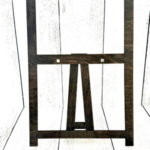 May include: A dark brown wooden easel stand with a rectangular frame. The easel is made of wood and has a simple design.