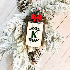 May include: A wooden Christmas ornament shaped like a sled with a white background and a green wreath with the letter K in the center. A red bow is attached to the top of the ornament.