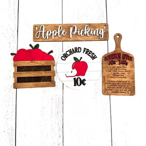 Puede incluir: Carteles de madera con tem&aacute;tica de recogida de manzanas. Incluye un letrero que dice "Apple Picking", una caja de manzanas rojas, un letrero que dice "Orchard Fresh 10&cent;" y una tabla de cortar con una receta de Bourbon Apple Hot Toddy.