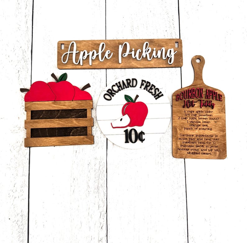Puede incluir: Carteles de madera con tem&aacute;tica de recogida de manzanas. Incluye un letrero que dice "Apple Picking", una caja de manzanas rojas, un letrero que dice "Orchard Fresh 10&cent;" y una tabla de cortar con una receta de Bourbon Apple Hot Toddy.