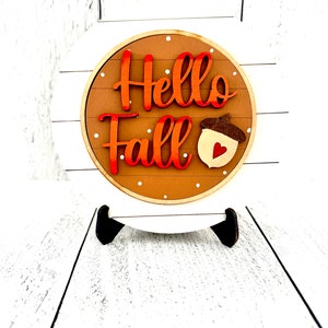 May include: A circular wooden sign featuring the text Hello Fall in orange and red gradient lettering. The sign has a brown background with white polka dots and a small wooden acorn icon with a red heart cutout, mounted on a white shiplap base.