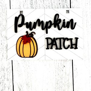 May include: A white wooden sign with black lettering that reads "Pumpkin Patch". A small orange pumpkin with a brown stem and a red leaf is on the left side of the sign.