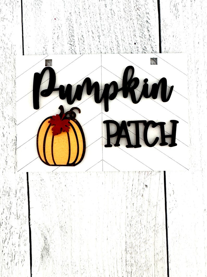 May include: A white wooden sign with black lettering that reads "Pumpkin Patch". A small orange pumpkin with a brown stem and a red leaf is on the left side of the sign.