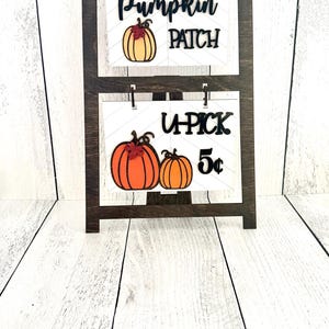 May include: A wooden stand with two signs. The top sign says "Pumpkin Patch" with a small orange pumpkin graphic. The bottom sign says "U-Pick 5&cent;" with two small orange pumpkins.