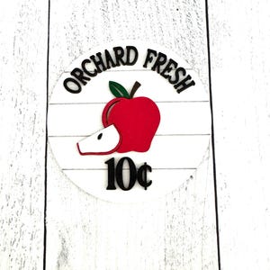 Puede incluir: Un letrero redondo de madera blanca con las palabras "ORCHARD FRESH" en negro, arqueadas sobre un gr&aacute;fico de manzana roja con una hoja verde y una rodaja. El precio "10&cent;" est&aacute; debajo de la manzana. El letrero est&aacute; sobre un fondo de madera blanca.