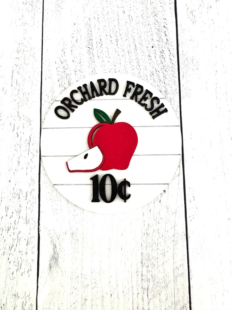 Puede incluir: Un letrero redondo de madera blanca con las palabras "ORCHARD FRESH" en negro, arqueadas sobre un gr&aacute;fico de manzana roja con una hoja verde y una rodaja. El precio "10&cent;" est&aacute; debajo de la manzana. El letrero est&aacute; sobre un fondo de madera blanca.