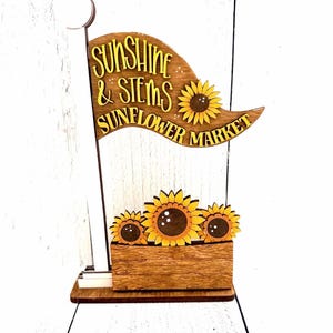 May include: A wooden decorative sign featuring a flag shape with the text "SUNSHINE & STEMS SUNFLOWER MARKET" in yellow lettering. The design includes three yellow sunflowers and a rectangular wooden base with a wood grain texture.