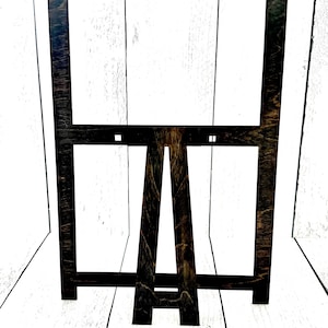 May include: A black wooden easel with a rectangular frame. The easel is made of wood and has a dark brown stain. The frame is made of wood and has a black stain.