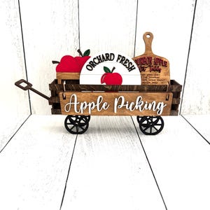 Puede incluir: Un carro de madera para recoger manzanas con ruedas negras, que muestra el texto "Apple Picking" en escritura blanca. Incluye un letrero que dice "Orchard Fresh" y una tabla de cortar de madera con texto. Tambi&eacute;n se ven manzanas rojas.