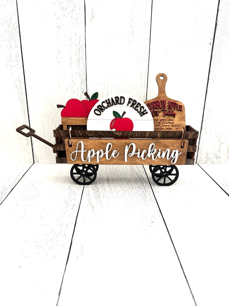 Puede incluir: Un carro de madera para recoger manzanas con ruedas negras, que muestra el texto "Apple Picking" en escritura blanca. Incluye un letrero que dice "Orchard Fresh" y una tabla de cortar de madera con texto. Tambi&eacute;n se ven manzanas rojas.
