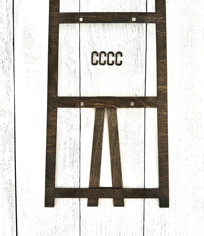 May include: A dark brown wooden easel with a rectangular frame. The frame has a white background and the text "CCCC" is printed in black.
