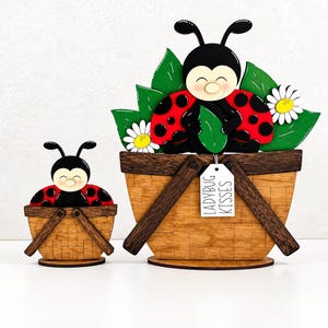 May include: Two wooden decorative items shaped like baskets with ladybugs, green leaves, and white daisies. The larger basket features a white rectangular tag hanging from the rim with the black printed text LADYBUG KISSES.