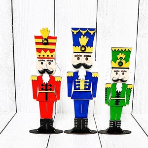 May include: Three flat wooden nutcracker figures standing in a row. The left figure wears a red uniform, the center figure wears a blue uniform, and the right figure wears a green uniform. Each nutcracker has a black mustache, white beard, and hat.