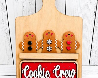 May include: A light-colored wooden cutting board shaped sign featuring three gingerbread figures holding hands. The figures have heart-shaped cheeks and different buttons. Below them is a red rectangular sign with the text Cookie Crew.