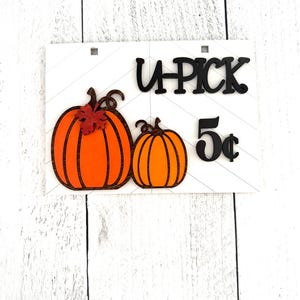 May include: A white wooden sign with black lettering that reads "U-PICK 5&cent;". There are two orange pumpkins with brown stems and a small red leaf on the larger pumpkin.
