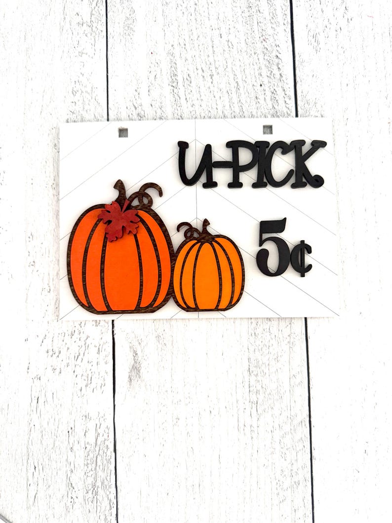 May include: A white wooden sign with black lettering that reads "U-PICK 5&cent;". There are two orange pumpkins with brown stems and a small red leaf on the larger pumpkin.