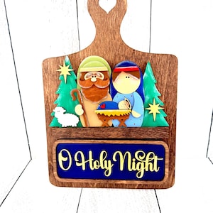 May include: A wooden decorative board with a heart-shaped cutout at the top, featuring a layered nativity scene with figures, green trees, and a white sheep. Below the scene, a blue rectangular plaque displays the yellow text O Holy Night.
