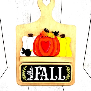 May include: Wooden fall-themed decorative cutting board with a heart-shaped handle. Features three pumpkins in white, orange, and yellow, with a black bird on each side. The words "hello FALL" are displayed on a black chalkboard.