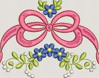 May include: An embroidery design featuring a central pink ribbon tied in a bow, flanked by clusters of blue flowers with yellow centers, green leaves, and small white pearl-like accents arranged in a symmetrical, decorative horizontal pattern.