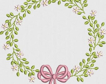 May include: A floral wreath design featuring a circle of green leaves and small white flowers. A pink bow is at the bottom center. The design is embroidered on a white background.
