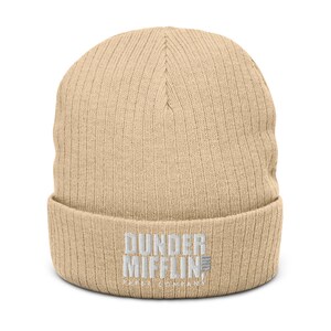 May include: A beige knitted beanie with the text "Dunder Mifflin Paper Company" embroidered in white on the front.