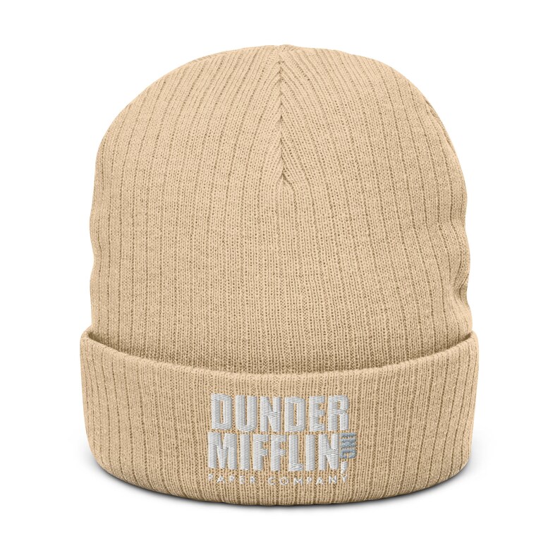 May include: A beige knitted beanie with the text "Dunder Mifflin Paper Company" embroidered in white on the front.