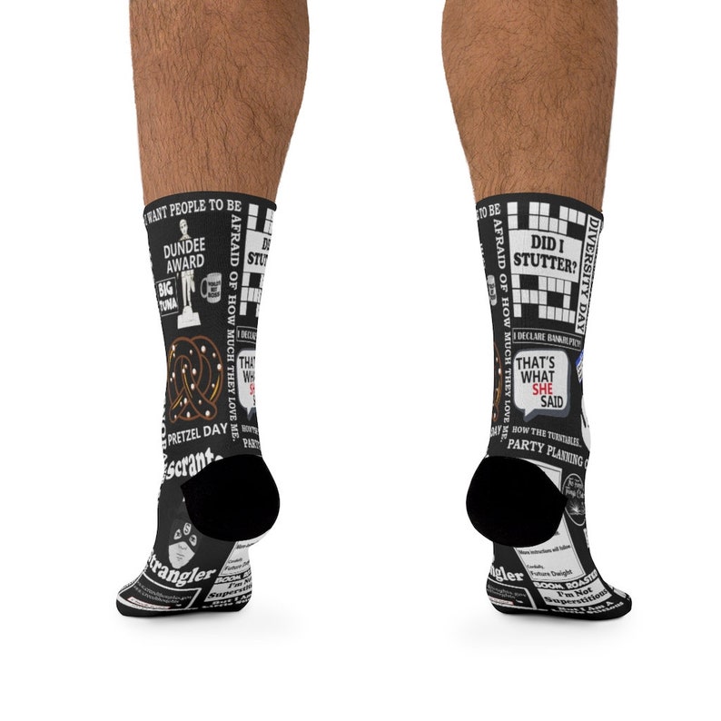 May include: A pair of black socks with a white design featuring quotes and crossword puzzles from the popular TV show "The Office". The socks feature the text "Dundee Award", "Pretzel Day", "That's What She Said", "Did I Stutter?" and "Diversity Day".