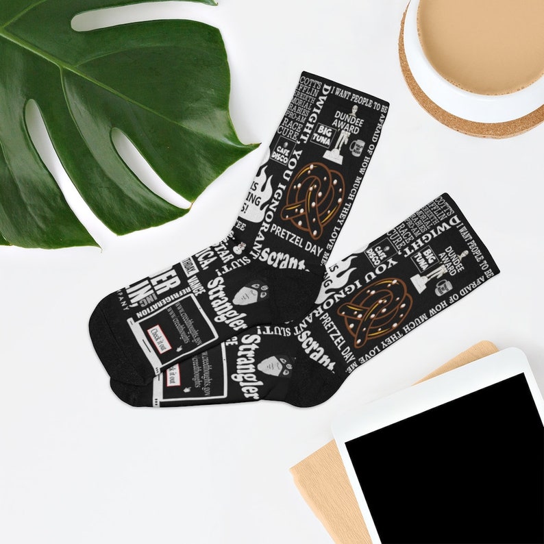May include: Black and white socks with a pattern of text and images from the TV show "The Office". The text includes quotes from the show, such as "I want people to be afraid of how much they love me" and "Pretzel Day". The images include a pretzel, a Dundie Award, and a big tuna.