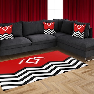 May include: A red and black area rug with a white zig-zag pattern and a white symbol of interlocking triangles. The rug is in front of a gray sectional sofa with three black and red pillows featuring the same symbol.