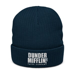 May include: A navy blue knitted beanie with a white embroidered logo that reads "Dunder Mifflin, Inc. Paper Company."