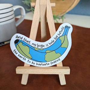 May include: A sticker in the shape of a banana, with a blue and green design resembling a world map. The sticker is displayed on a small wooden easel. Text on the sticker reads, "And that, my leige, is how we know the earth to be banana shaped."
