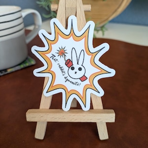 May include: A white sticker with a cartoon rabbit holding a dynamite stick, set against a yellow and orange burst design. The text "That rabbit's dynamite!" is printed on the sticker. The sticker is displayed on a small wooden easel.