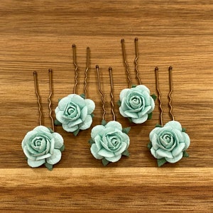 May include: Six mint green fabric rose hair clips with gold bobby pins. The hair clips are arranged in a row on a wooden surface.