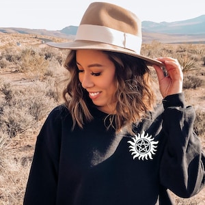 Może przedstawiać: Czarna bluza z białym symbolem z przodu. Symbol to pięcioramienna gwiazda w okręgu z promieniami wychodzącymi z okręgu. Osoba nosząca bluzę ma na sobie brązowy kapelusz fedora.