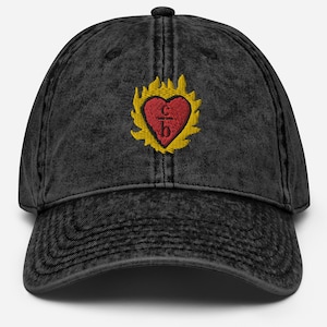 Op de afbeelding: Een zwarte vintage gewassen denim baseballcap met een geborduurd rood hart met een kruis en de letters 'IHS' erin. Het hart is omgeven door gele vlammen.