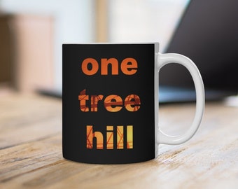 Op de afbeelding: Een zwarte keramische mok met de woorden "one tree hill" in oranje en gele letters. De mok heeft een wit handvat en staat op een houten oppervlak. Een laptop is zichtbaar op de achtergrond.