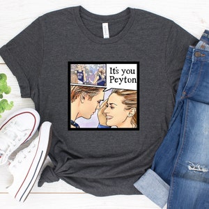 May include: A dark gray t-shirt with a comic book style image of a couple looking at each other. The speech bubble says "It's you Peyton".