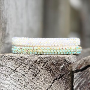 Könnte beinhalten: Zwei Perlenarmbänder, eines weiß und eines irisierend, liegen auf einer verwitterten Holzoberfläche. Die Armbänder bestehen aus kleinen, runden Perlen, wodurch eine strukturierte Optik entsteht. Das irisierende Armband hat einen dezenten Grün- und Blauton.