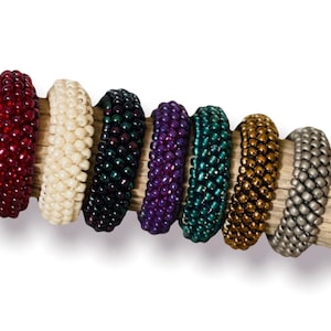 Könnte beinhalten: Sieben kreisförmige Ringe aus kleinen, gleichmäßigen Perlen in verschiedenen Farben, darunter Dunkelrot, Creme, Dunkelblau, Violett, Petrol, Metallic-Gold und mattes Silber. Jeder Ring weist eine dichte, gewebte Textur aus einzelnen Perlen auf.