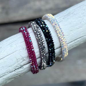 May include: Four beaded bracelets in pink, silver, black, and iridescent white. The bracelets are made of small, sparkling beads and are arranged on a white wooden surface. The bracelets are a fashion accessory.