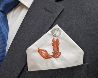 May include: A white pocket square featuring an embroidered orange fox sitting with its tail curled, positioned below a circular silver-toned embroidered moon, tucked into the breast pocket of a dark charcoal suit jacket with a blue tie.