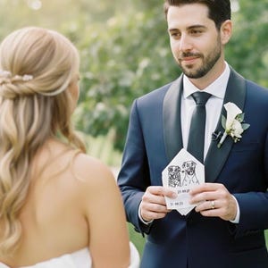 Groom in a navy suit and white boutonniere smiling at the bride while holding a pocket square embroidered with two dog portraits and a date, given as a wedding day gift.