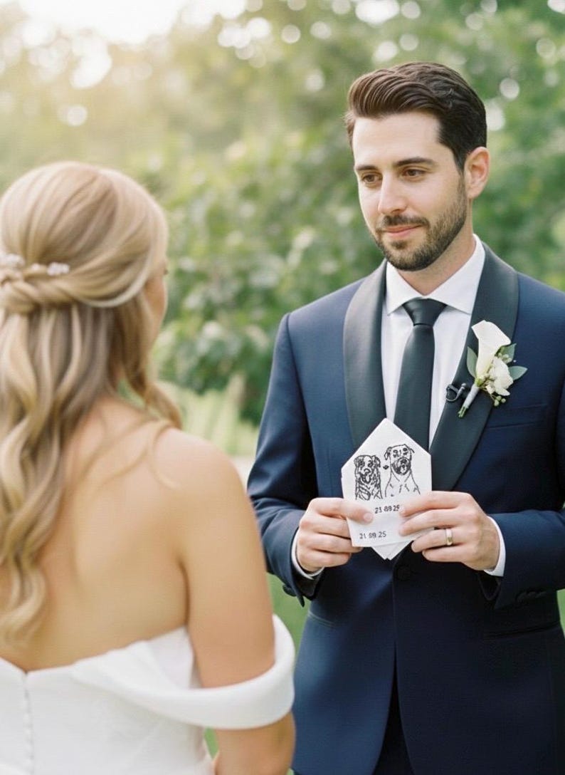 Groom in a navy suit and white boutonniere smiling at the bride while holding a pocket square embroidered with two dog portraits and a date, given as a wedding day gift.