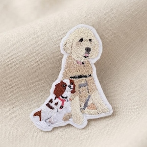 May include: An embroidered patch featuring a large, light tan, curly-haired dog sitting upright next to a smaller, white and brown Cavalier King Charles Spaniel. The patch has a white border and detailed stitching depicting the two dogs.