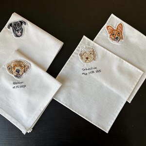Four white embroidered handkerchiefs with custom dog portraits (a black Labrador, a golden dog, a fluffy doodle and a corgi), some with a name and date stitched below.