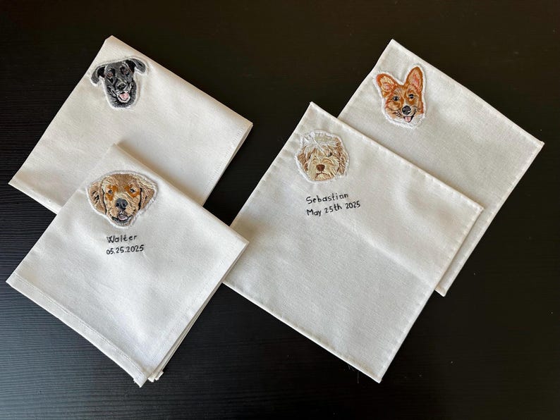 Four white embroidered handkerchiefs with custom dog portraits (a black Labrador, a golden dog, a fluffy doodle and a corgi), some with a name and date stitched below.