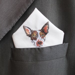White cotton pocket square with a hand-embroidered portrait of a brown and white dog with large ears, folded in the breast pocket of a charcoal gray suit jacket.