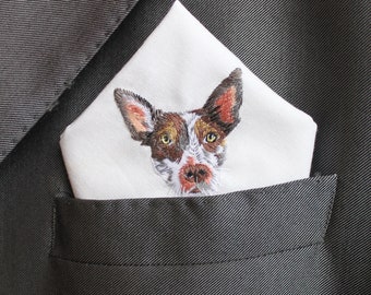 May include: A white pocket square with an embroidered dog's face, in brown, white, and black, sits in the pocket of a dark grey suit jacket. The dog's face is the focal point of the square.