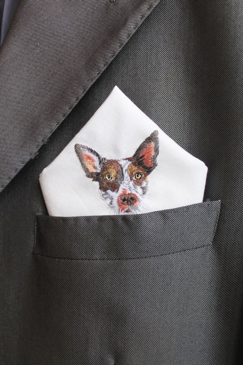 White cotton pocket square with a hand-embroidered portrait of a brown and white dog with large ears, folded in the breast pocket of a charcoal gray suit jacket.